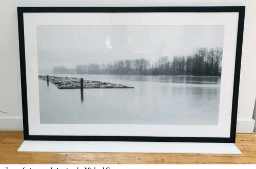 Large black-framed black-and-white photograph of a calm lake with bare trees along the far shore, leaning against a white wall on a wooden floor.