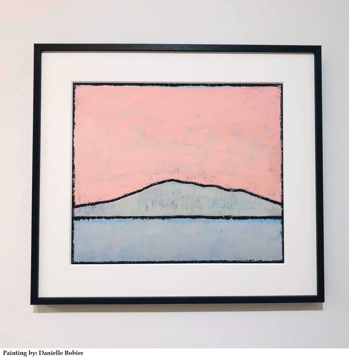 Framed abstract landscape painting: pink sky, dark ridge, and pale blue water on a white wall.