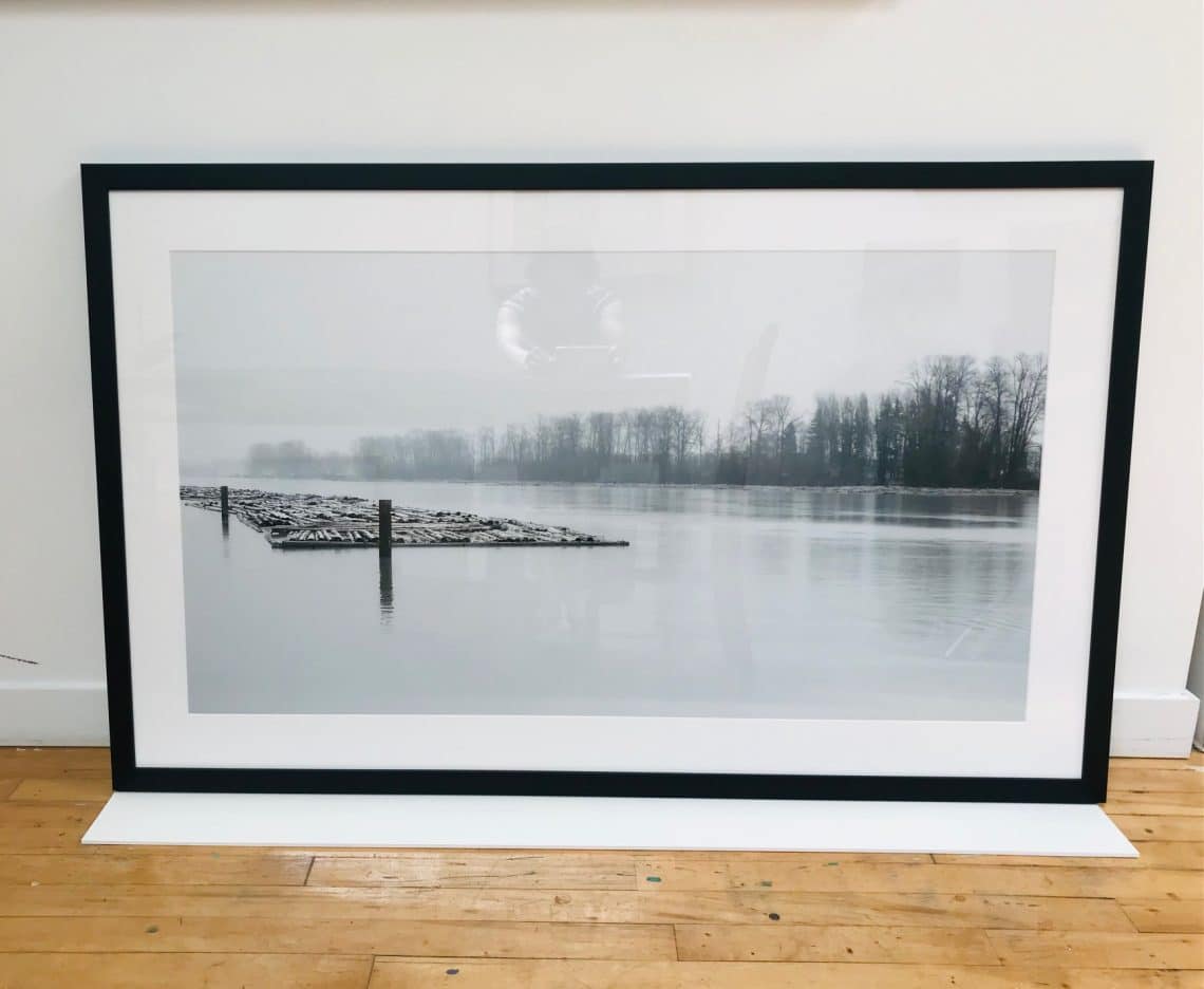 Framed black-and-white photograph of a calm lake with floating logs and a line of bare trees in the distance.