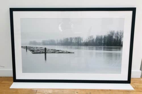 Framed black-and-white photograph of a calm lake with floating logs and a line of bare trees in the distance.