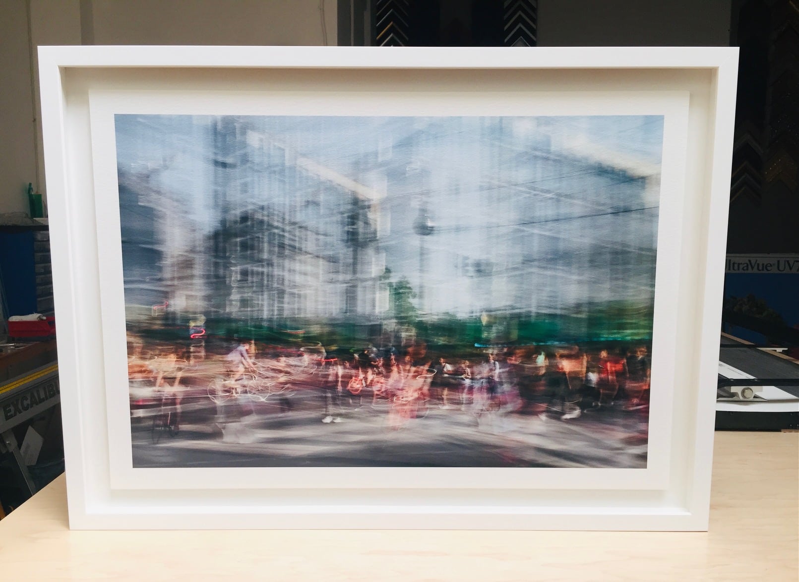 Framed print of a crowded city street with blurred pedestrians and bicycles in motion under a blue sky, captured with long exposure.